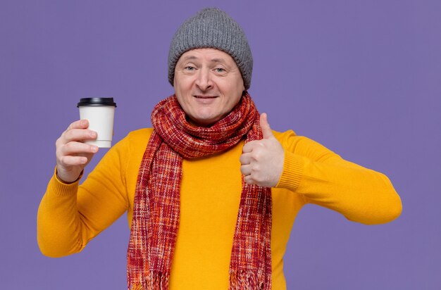 Smiling adult man with winter hat and scarf around his neck holding paper cup and thumbing up 