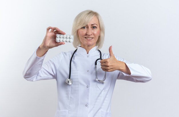 Smiling adult slavic female doctor in medical robe with stethoscope holding medicine tablet in blister pack and thumbing up isolated on white background with copy space