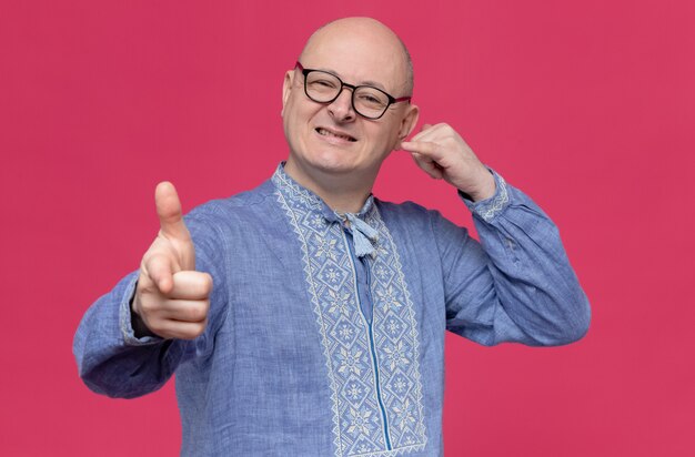 Smiling adult slavic man in blue shirt wearing optical glasses gesturing call me sign and pointing at front