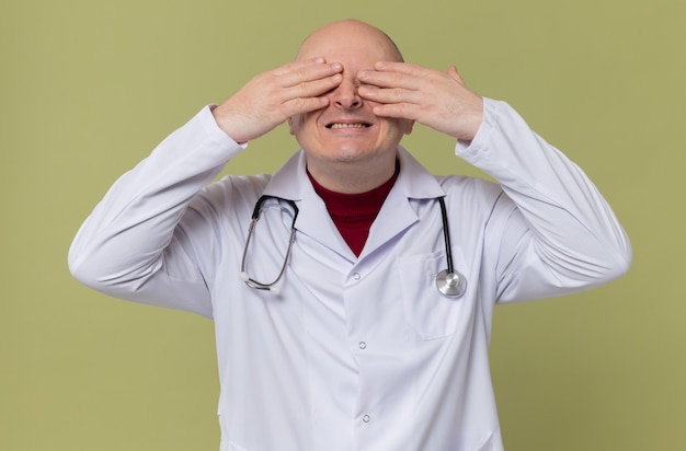 Smiling adult slavic man in doctor uniform with stethoscope closing his eyes with hands