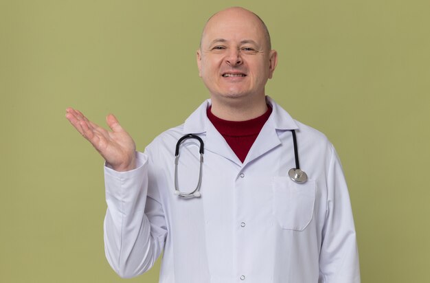 Smiling adult slavic man in doctor uniform with stethoscope keeping open his hand 