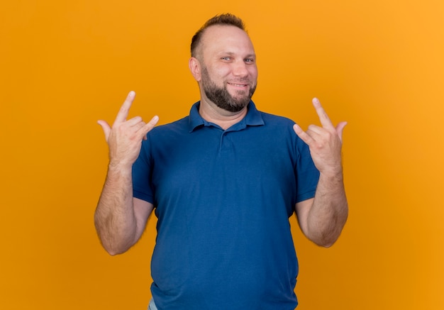 Smiling adult slavic man doing rock signs isolated on orange wall