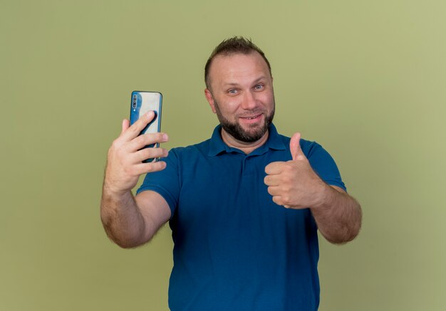 Smiling adult slavic man holding mobile phone and showing thumb up 