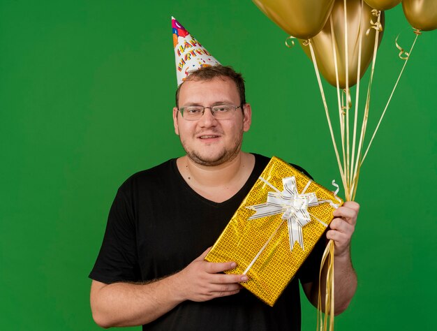 Smiling adult slavic man in optical glasses wearing birthday cap holds helium balloons and gift box 