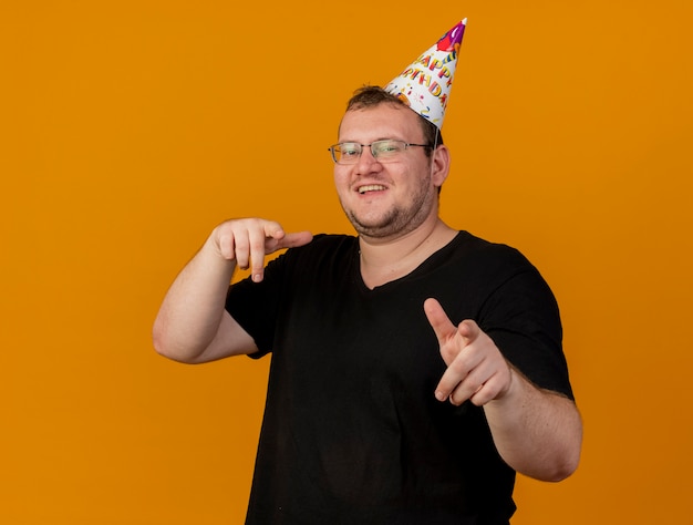 Smiling adult slavic man in optical glasses wearing birthday cap looks and points at camera 