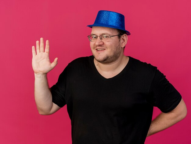 파란색 파티 모자를 쓰고 광학 안경에 웃는 성인 슬라브 남자는 제기 손으로 서있다.
