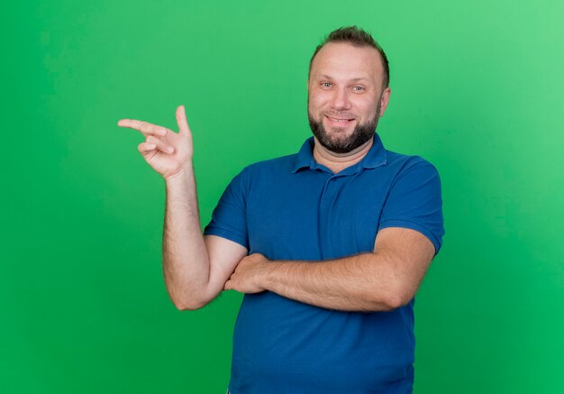 Smiling adult slavic man  pointing at side isolated on green wall