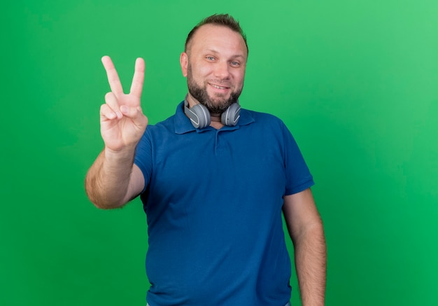 Улыбающийся взрослый славянский мужчина в наушниках на шее смотрит на знак мира на зеленой стене с копией пространства