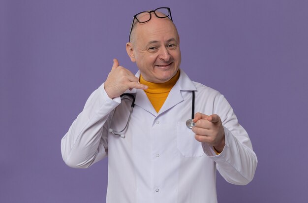 Smiling adult slavic man with optical glasses in doctor uniform with stethoscope gesturing call me sign and looking at front