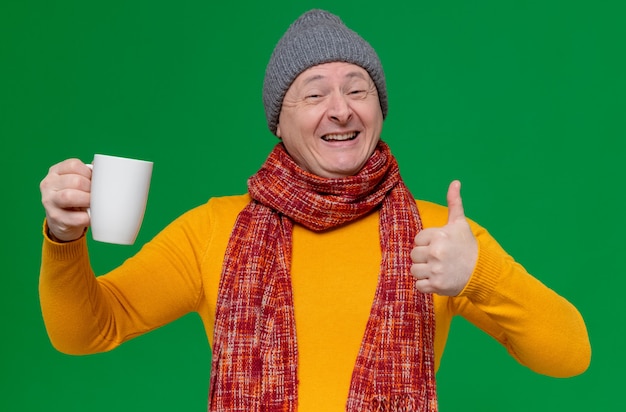 Smiling adult slavic man with winter hat and scarf around his neck holding cup and thumbing up