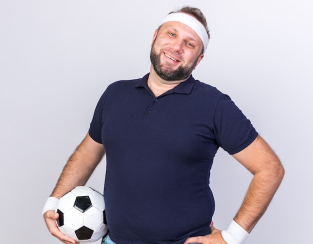 コピースペースで白い壁に分離されたボールを保持しているヘッドバンドとリストバンドを身に着けている大人のスラブスポーティーな男の笑顔