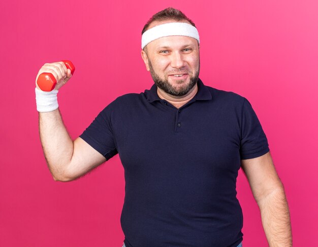 smiling adult slavic sporty man wearing headband and wristbands holding dumbbell isolated on pink wall with copy space