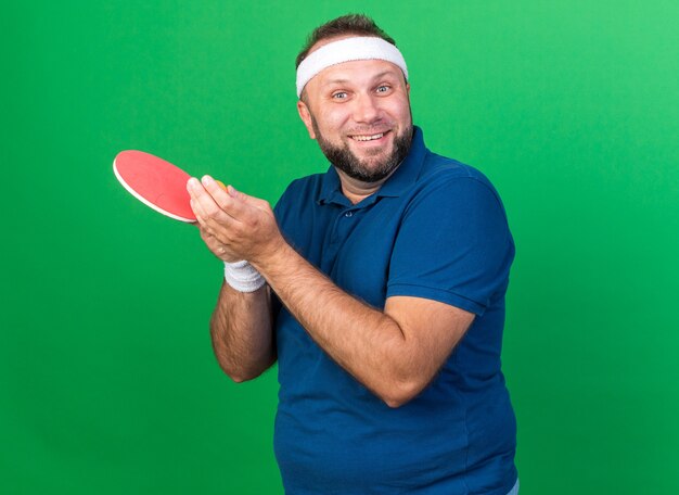 smiling adult slavic sporty man wearing headband and wristbands holding ping-pong ball and racket isolated on green wall with copy space