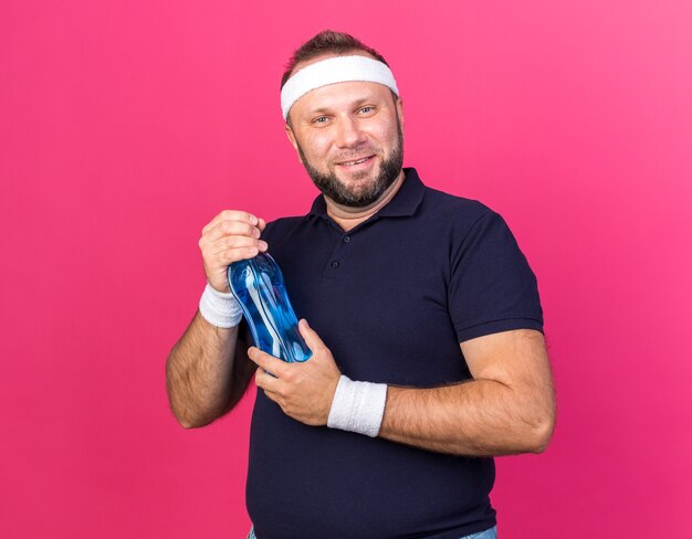 smiling adult slavic sporty man wearing headband and wristbands holding water bottle isolated on pink wall with copy space