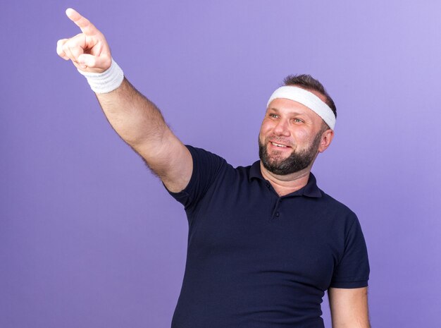 smiling adult slavic sporty man wearing headband and wristbands looking and pointing up isolated on purple wall with copy space