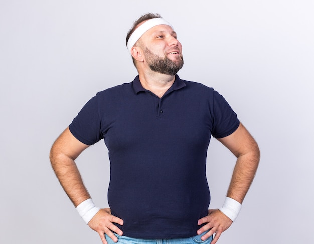 smiling adult slavic sporty man wearing headband and wristbands putting hands on waist and looking at side isolated on white wall with copy space