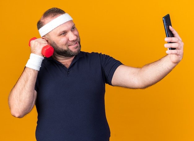 smiling adult slavic sporty man wearing headband and wristbands taking selfie holding dumbbell isolated on orange wall with copy space