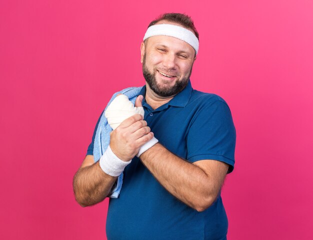 smiling adult slavic sporty man with towel on shoulder wearing headband and wristbands holding his hand isolated on pink wall with copy space