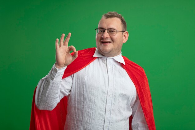 Smiling adult slavic superhero man in red cape wearing glasses  doing ok sign isolated on green wall with copy space