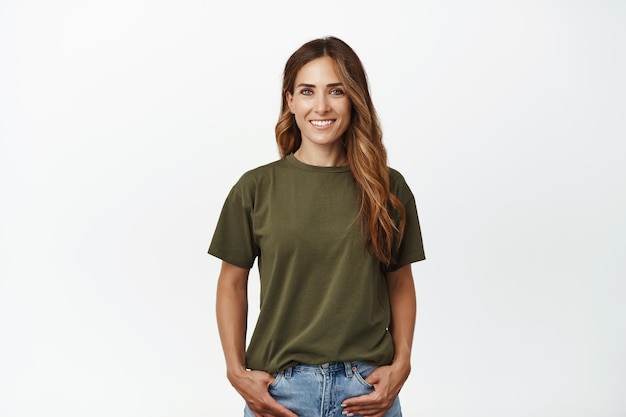 Smiling adult woman in casual clothes, holding hands in pockets, smiling pleased and confident, standing against white background