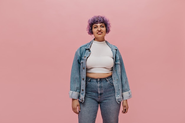 Smiling adult woman with short curly purple hair in white top, in denim jacket and tight jeans posing.