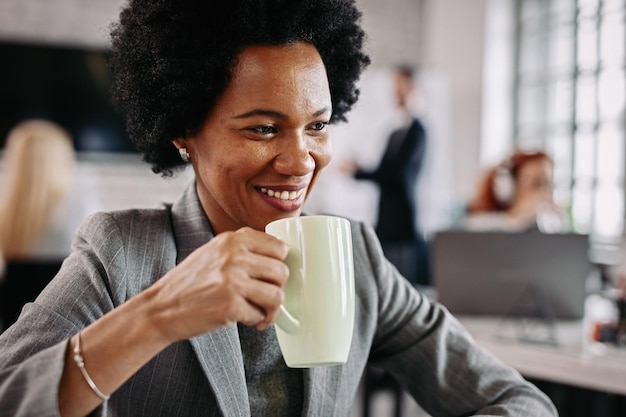 Sorridente imprenditrice afroamericana che beve caffè mentre è al lavoro ci sono persone in background