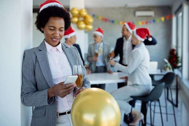 사무실에서 크리스마스 파티를 하는 동안 웃고 있는 아프리카계 미국인 여성 사업가가 휴대전화로 문자를 보냈다