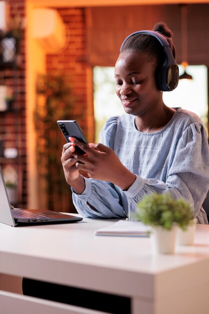 휴대 전화에 메시지를 입력 하는 웃는 아프리카계 미국인 사업가. 직장에서 휴식을 취하고 현대적인 홈 오피스에서 스마트폰으로 재미있는 비디오를 보는 여성 프리랜서