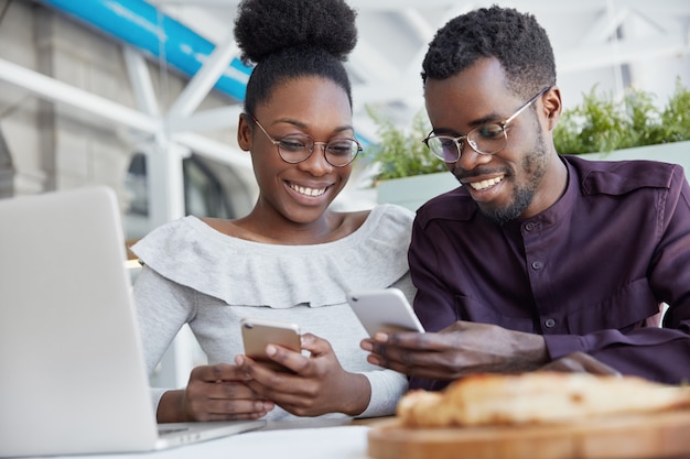Smiling African American friends meet together at cafe, use modern technologies for entertainment. Dark skinned delighted young female and male hold smart phones