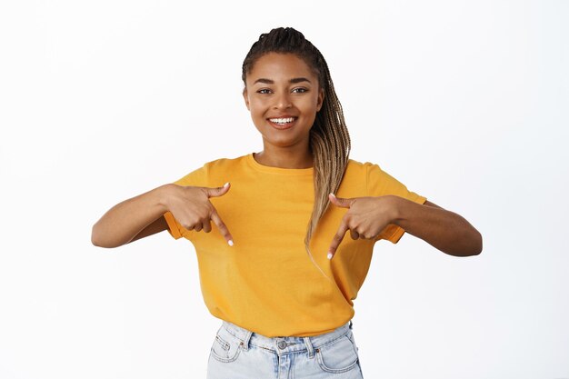 Smiling african american girl pointing fingers down with proud and confident face showing advertisement standing in yellow tshirt over white background