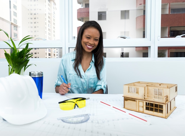 Smiling African American lady on chair with pen near safety helmet and model of house on table