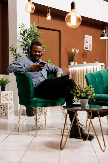 Smiling African American man wearing wireless headphones relaxing in hotel lobby using smartphone watching online video. Black guy browsing internet while sitting in lounge area at luxury resort