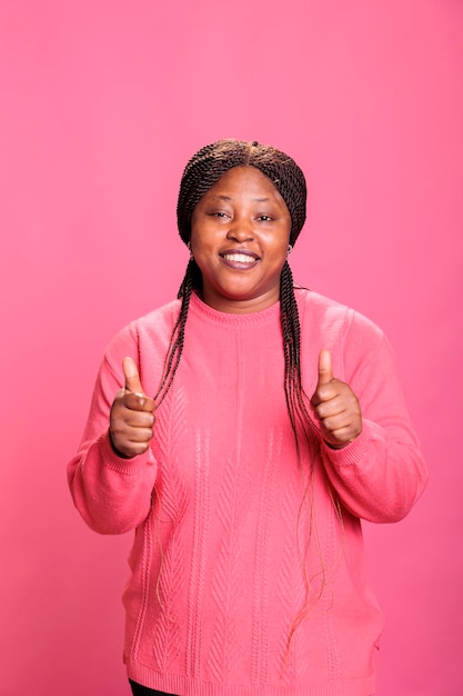 Free photo smiling african american model showing thumbs up sign standing over pink background in studio. happy cheerful woman with joyful facial expression giving approval gesture, person doing like sign