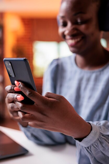 携帯電話を保持しているヘッドフォンでアフリカ系アメリカ人の女性の笑顔。仕事で休憩し、現代のホームオフィスでスマートフォンで面白いビデオを見ている女性のSMMマネージャー