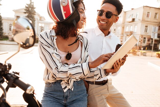 Smiling african couple sitting on modern motorbike and using tablet computer on the street while looking to each other