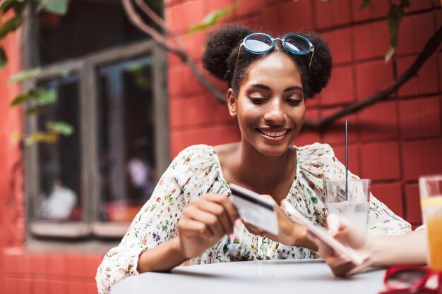 Smiling african girl in blouse and sunglasses on head happily holding credit card while spending time in cozy courtyard of cafe