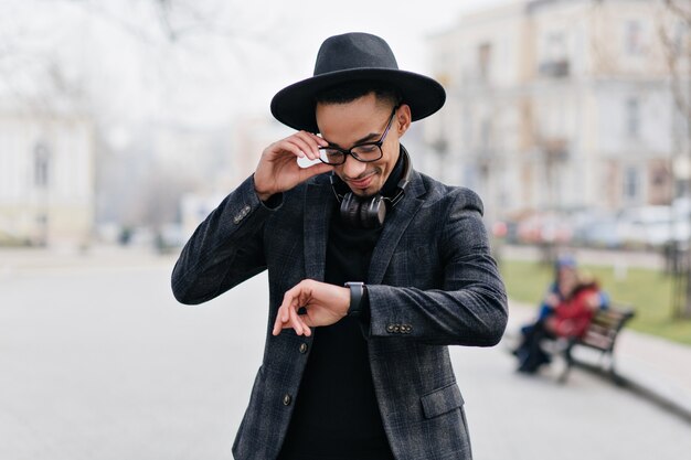 Smiling african guy looking at wristwatch while standing on the street. Outdoor portrait of happy young man waiting girlfriend in park.