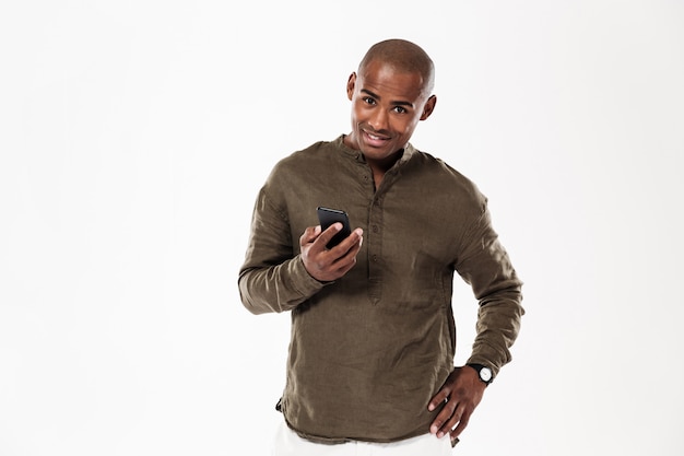 Smiling african man holding arm on hip and using smartphone while looking