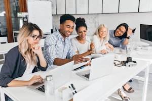 Smiling african student pointing with pencil at laptop screen. concentrated blonde woman in glasses propping chin with hand while working with computer in office.