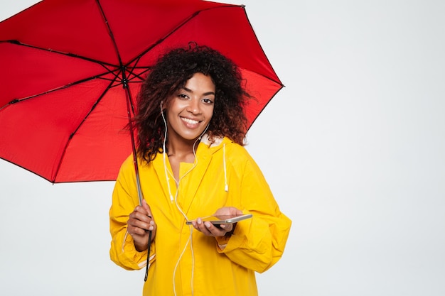 Donna africana sorridente in impermeabile che si nasconde sotto l'ombrello e la musica d'ascolto