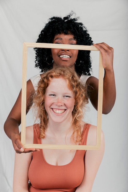 Smiling african young woman holding wooden frame in front of caucasian female