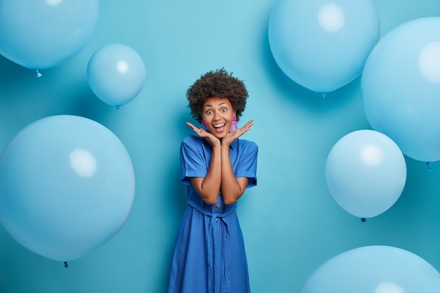웃는 아프리카 계 미국인 소녀는 손바닥을 얼굴에 펼치고 멋진 여름 파티를 즐기고 긴 파란색 유행의 드레스를 입고 팽창 된 풍선 위에 포즈를 취하고 행복한 분위기에 있습니다. 축하 및 라이프 스타일 컨셉