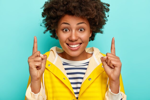 Smiling Afro American woman with happy expression, shows direction above, wears striped jumper and yellow raincoat, isolated over blue background.