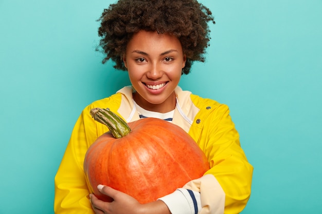 Smiling Afro woman holds pumpkin in fall,  wears yellow protective raincoat, has happy mood, stands against blue background.