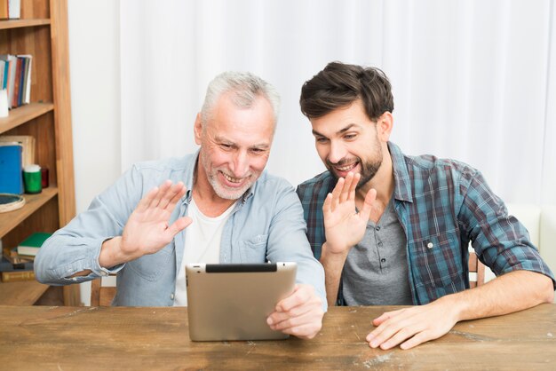 Smiling aged man and happy young guy waving hands and using tablet at table