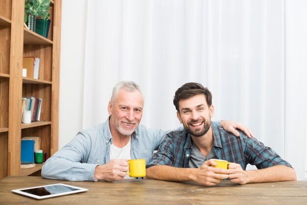 Free photo smiling aged man hugging happy young guy with cups at table with tablet