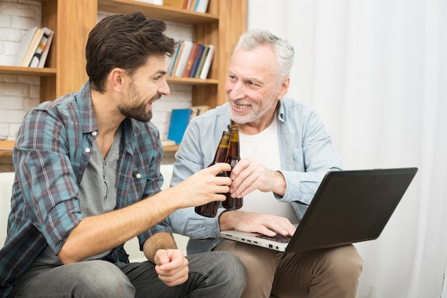 Smiling aged man and young guy clanging bottles and using laptop on sofa