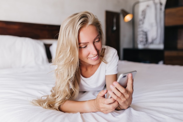 Foto gratuita sorridente attraente donna sdraiata a letto con il telefono. splendida donna bionda agghiacciante in camera da letto e messaggio di testo.