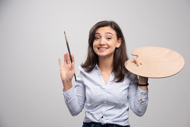 Smiling artist holding paintbrush and palette over gray wall. 