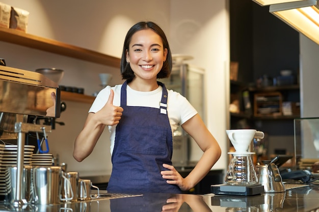 無料写真 カフェの制服を着た笑顔のアジア人バリスタの女の子が、コーヒーに新しい承認を勧めるokのサインを示している